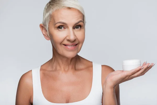 Happy senior woman showing container with cosmetic cream isolated on grey — Stock Photo