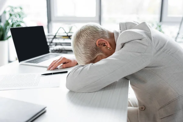 Müde Seniorin mit grauen Haaren liegt auf Schreibtisch neben Laptop — Stockfoto