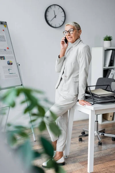Cheerful senior businesswoman in glasses talking on cellphone in office — Stock Photo