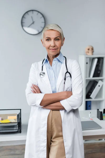 Pleased doctor with grey hair in white coat standing with crossed arms — Stock Photo