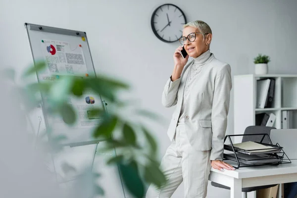 Souriant femme d'affaires senior dans des lunettes parler sur téléphone portable dans le bureau — Photo de stock