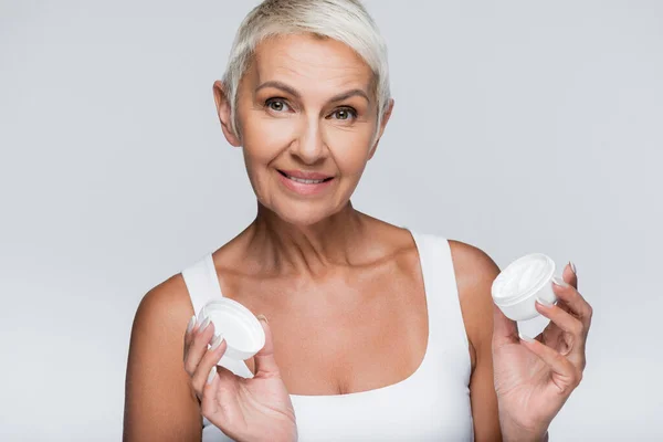 Happy elderly woman holding container with cosmetic cream isolated on grey — Stock Photo