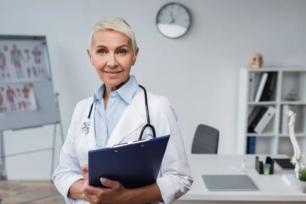 Alegre médico senior sosteniendo portapapeles y sonriendo en la clínica - foto de stock