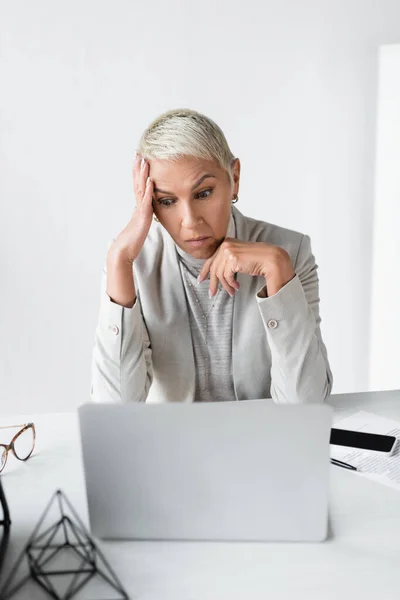 Femme d'affaires mécontente avec les cheveux gris regardant ordinateur portable près du smartphone sur le bureau — Photo de stock