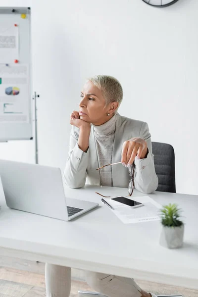 Pensive businesswoman with grey hair holding glasses at workplace — Stock Photo