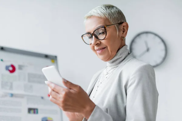 Pleased senior businesswoman in glasses looking at smartphone — Stock Photo
