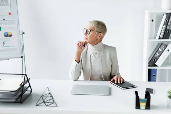 Pensativa mujer de negocios senior mirando lejos cerca de gadgets en el escritorio - foto de stock