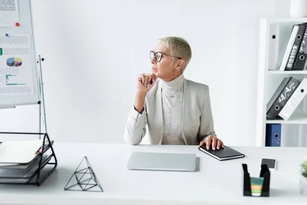 Pensive senior businesswoman looking away near gadgets on desk — Stock Photo