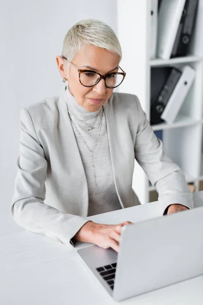 Empresária com cabelos grisalhos em óculos usando laptop no escritório — Fotografia de Stock