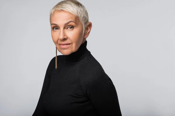 Donna anziana sorridente con orecchino lungo e dolcevita nero guardando la fotocamera isolata sul grigio — Foto stock