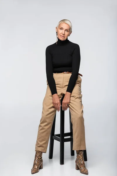 Volle Länge der stilvollen Seniorin in beiger Hose und Rollkragen posiert in der Nähe Stuhl auf grau — Stockfoto