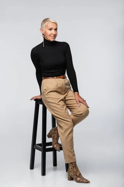 Full length of smiling senior woman in beige pants and black turtleneck posing near chair on grey — Stock Photo