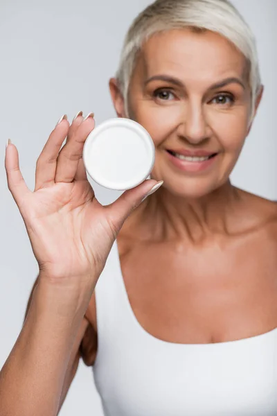 Mujer anciana positiva sosteniendo contenedor con crema cosmética aislada en gris - foto de stock