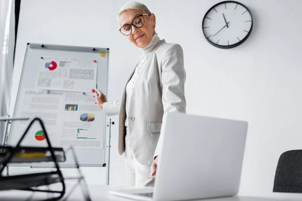 Smiling senior businesswoman in glasses looking at blurred laptop — Stock Photo