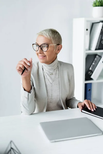 Mulher de negócios sênior feliz em óculos segurando caneta no escritório — Fotografia de Stock