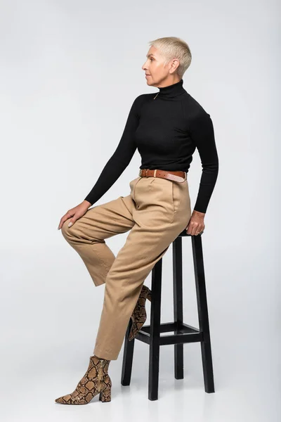 Longitud completa de la mujer mayor de moda en pantalones beige y cuello alto posando cerca de la silla en gris - foto de stock