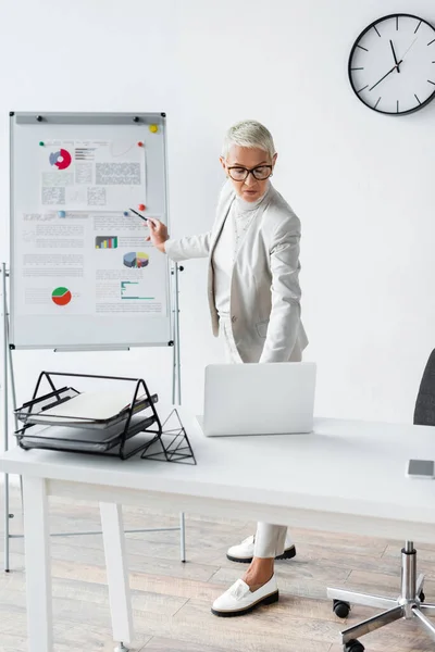 Старша бізнес-леді в окулярах дивиться на ноутбук, стоячи поруч з фліп-чартом — стокове фото