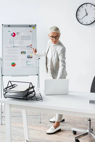 Senior businesswoman in glasses looking at laptop while standing near flip chart — Stock Photo