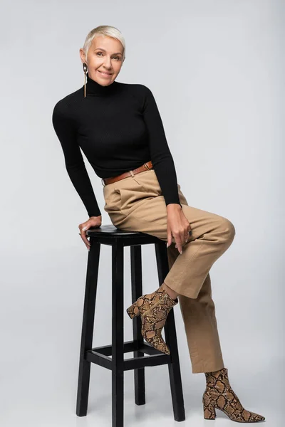Full length of cheerful senior woman in beige pants and turtleneck sitting on chair on grey — Stock Photo
