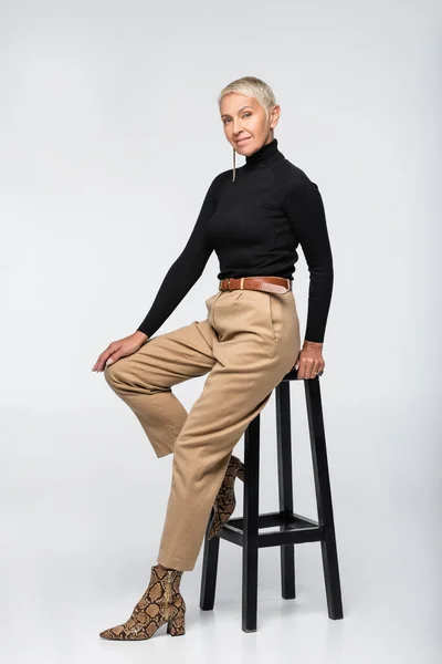 Full length of pleased senior woman in beige pants and turtleneck posing near chair on grey — Stock Photo