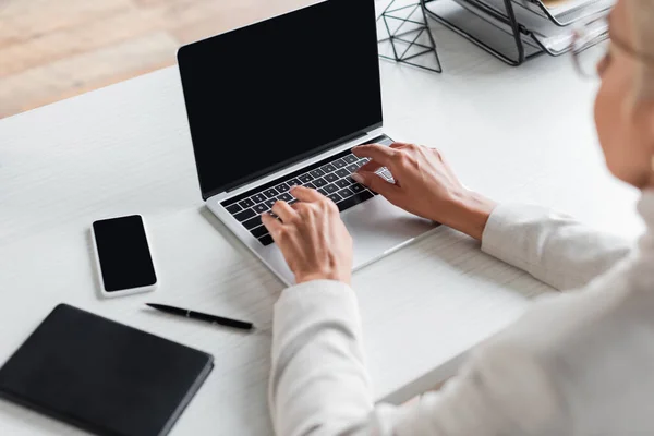 Mujer de negocios borrosa utilizando el ordenador portátil con pantalla en blanco cerca del teléfono inteligente en el escritorio - foto de stock