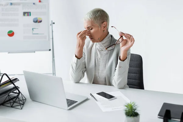 Müde Geschäftsfrau mit grauen Haaren, die Augen berührt und eine Brille in der Nähe des Laptops hält — Stockfoto