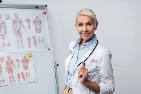 Happy senior doctor in white coat with stethoscope standing near flip chart with anatomical pictures — Stock Photo
