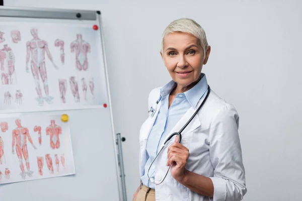 Heureux médecin aîné en manteau blanc avec stéthoscope debout près de flip chart avec des images anatomiques — Photo de stock