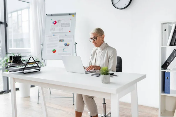 Donna d'affari anziana in occhiali usando il computer portatile in ufficio — Foto stock