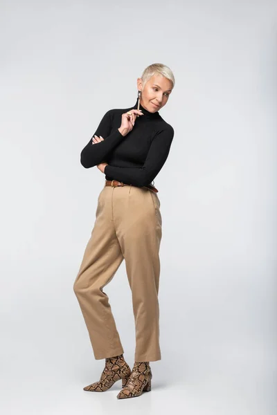 Longitud completa de mujer mayor complacida y elegante en pantalones beige y cuello alto posando en gris - foto de stock