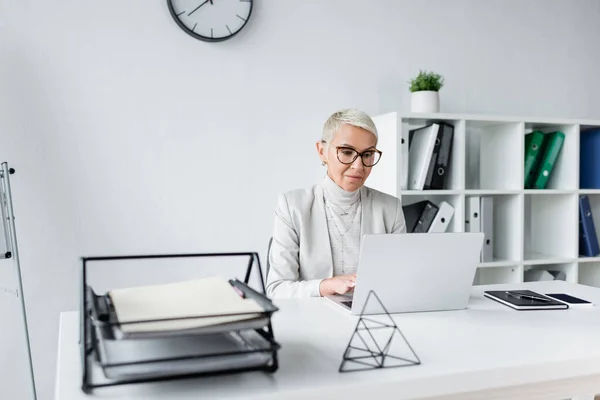 Donna d'affari con i capelli grigi in occhiali e tuta utilizzando il computer portatile in ufficio — Foto stock