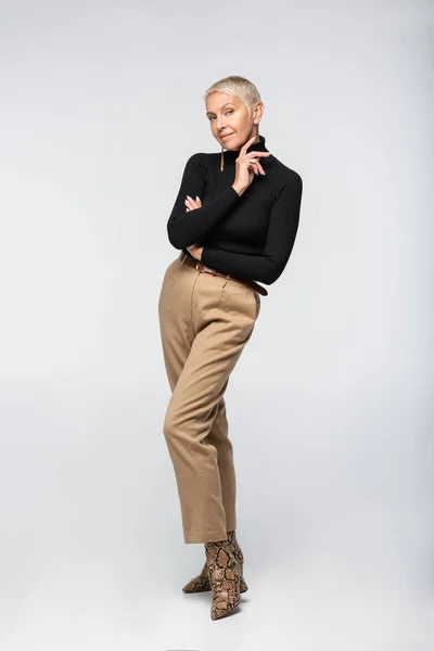 Volle Länge der zufriedenen und trendigen Seniorin in beiger Hose und Rollkragen posiert auf grau — Stockfoto