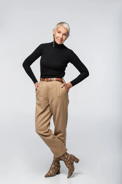 Full length of smiling senior woman in beige pants, black turtleneck and animal print boots posing with hands in pockets on grey — Stock Photo
