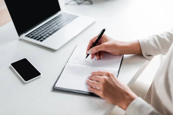 Vista parcial de la mujer de negocios escribiendo en cuaderno cerca de gadgets - foto de stock