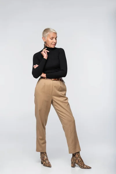 Full length of pleased and elegant senior woman in beige pants, black turtleneck and animal print boots posing on grey — Stock Photo