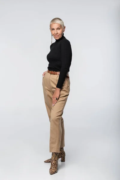 Full length of trendy senior woman in black turtleneck and animal print boots posing on grey — Stock Photo