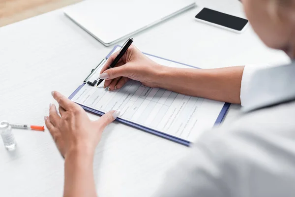 Cropped view of blurred doctor writing prescription near gadgets on desk — Stock Photo