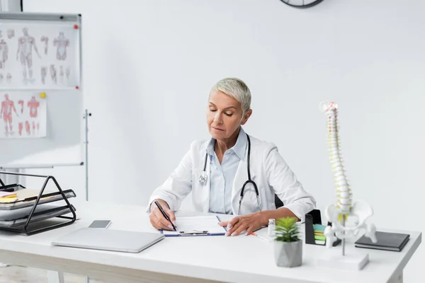 Arzt mit grauen Haaren schreibt Rezept in der Nähe von Geräten auf den Schreibtisch — Stockfoto