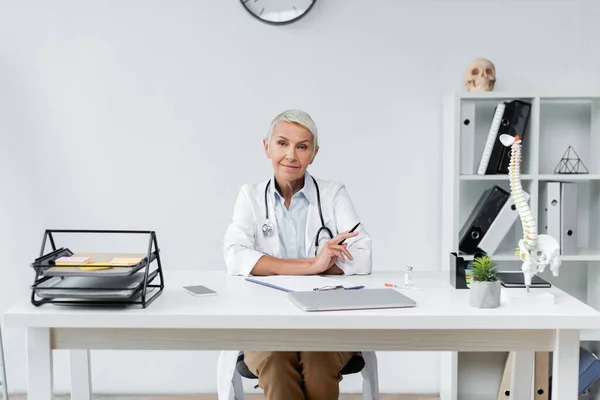 Alegre médico senior en bata blanca sonriendo cerca de dispositivos en el escritorio - foto de stock