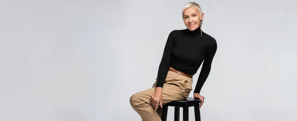 Zufriedene und trendige Seniorin in beiger Hose und Rollkragen sitzt auf Stuhl isoliert auf grau, Banner — Stockfoto