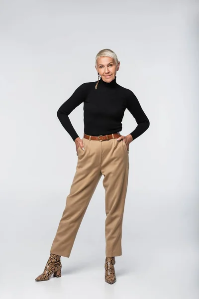 Full length of pleased and trendy senior woman in pants, black turtleneck and animal print boots posing with hands in pockets on grey — Stock Photo