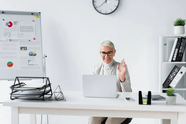 Sorridente empresária sênior acenando a mão durante videochamada no laptop no escritório moderno — Fotografia de Stock