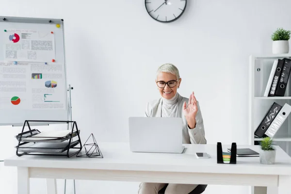 Lächelnde Seniorin winkt bei Videoanruf auf Laptop in modernem Büro — Stockfoto