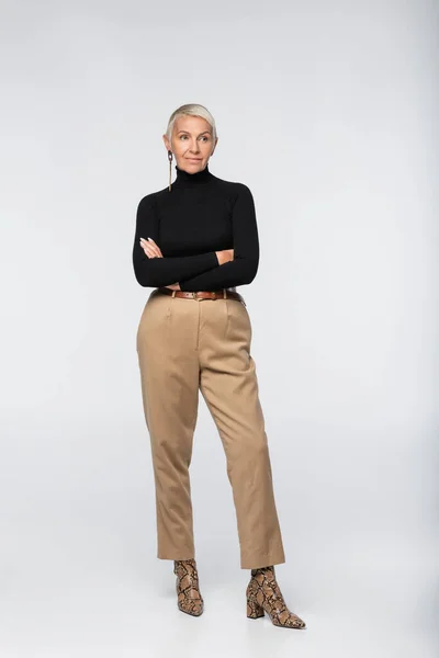Full length of trendy senior woman in beige pants, black turtleneck and animal print boots posing with crossed arms on grey — Stock Photo