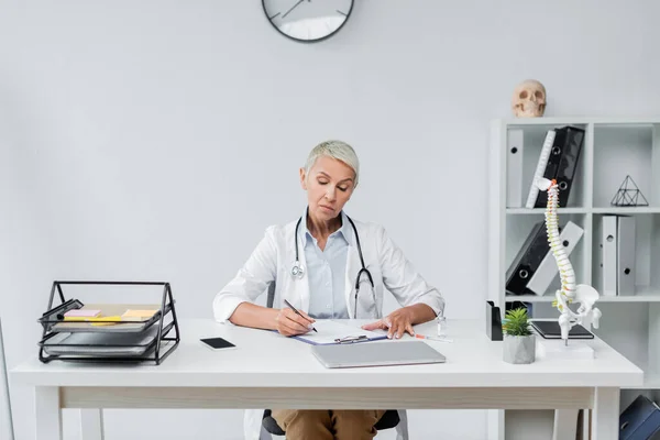 Médico senior en la prescripción de la bata blanca cerca de dispositivos en el escritorio - foto de stock