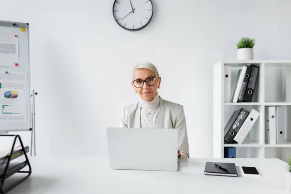 Senior businesswoman in glasses sitting at desk with devices in office — Stock Photo