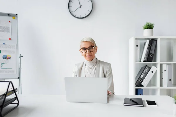 Empresária sênior em óculos sentados na mesa com dispositivos no escritório — Fotografia de Stock