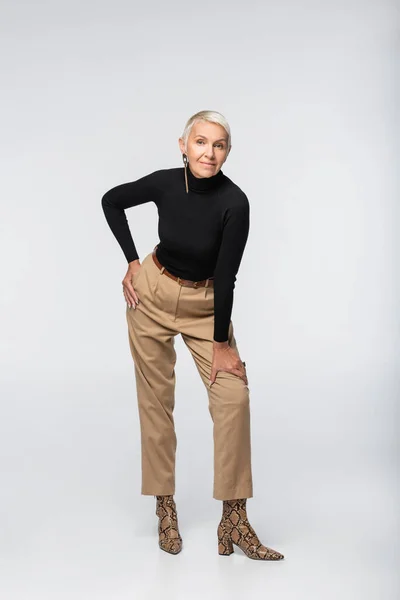Full length of pleased and trendy senior woman in beige pants, black turtleneck and animal print boots posing on grey — Stock Photo