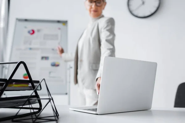 Laptop on desk near blurred businesswoman in office — Stock Photo