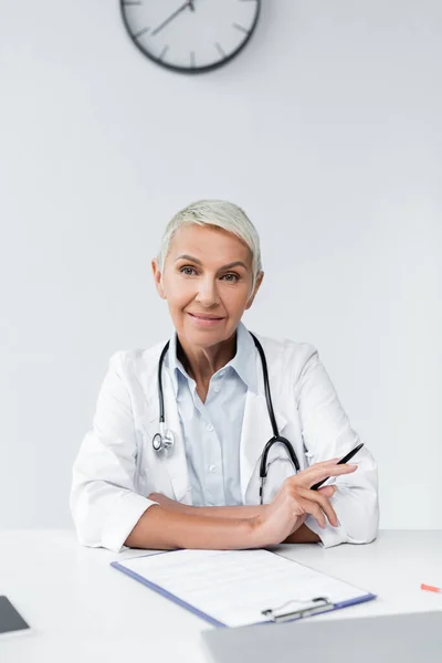 Happy senior doctor in white coat holding pen near clipboard on desk — Stock Photo